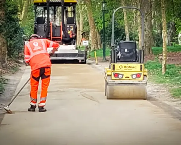 Un ouvrier prépare le chemin avec du sable stabilisé, tandis qu'un rouleau compresseur Bomag aplatit la surface.