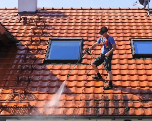 Un homme effectue le nettoyage toiture avec un nettoyeur haute pression sur un toit en tuiles rouges, sous un ciel bleu.