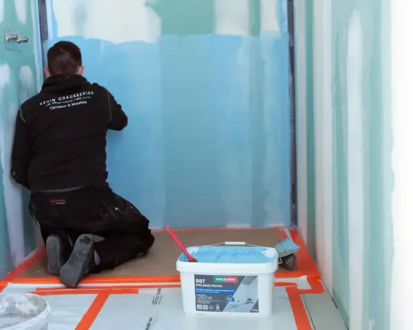 Un ouvrier applique un système d'étanchéité liquide bleu sur les murs d'une douche. Un seau de produit est visible au premier plan.