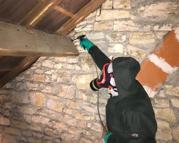 Personne en combinaison et masque effectuant un traitement de charpente par pulvérisation dans un grenier en pierre.