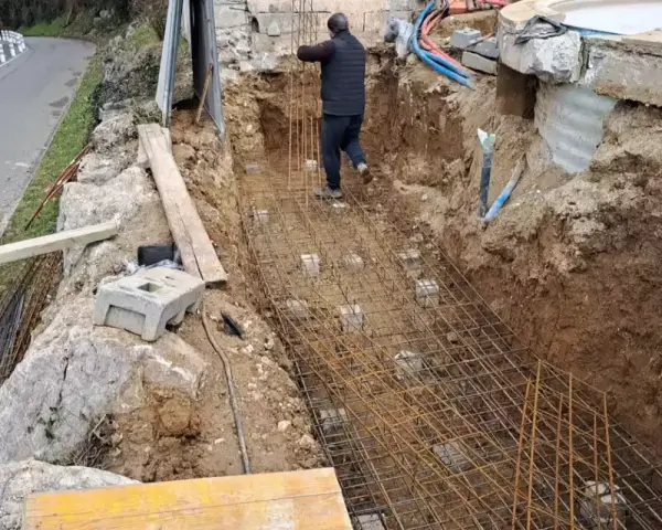 Un ouvrier travaille sur le ferraillage d'une fondation. Des armatures métalliques sont posées dans une fouille creusée dans la terre.