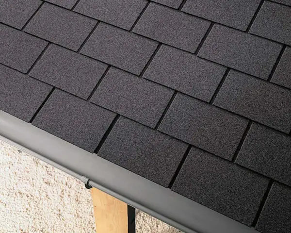 Détail d'une toiture en pose de bardeau bitumé gris foncé, avec une gouttière grise.