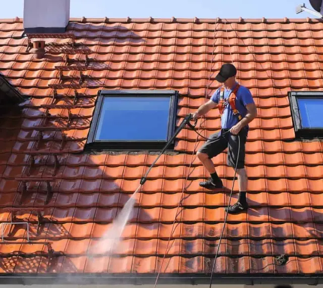 Un homme effectue le nettoyage toiture avec un nettoyeur haute pression sur un toit en tuiles rouges, sous un ciel bleu.