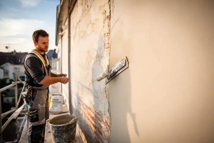 Un ouvrier applique un enduit sur agglo sur un mur extérieur, à l'aide d'une truelle. Le soleil se couche.