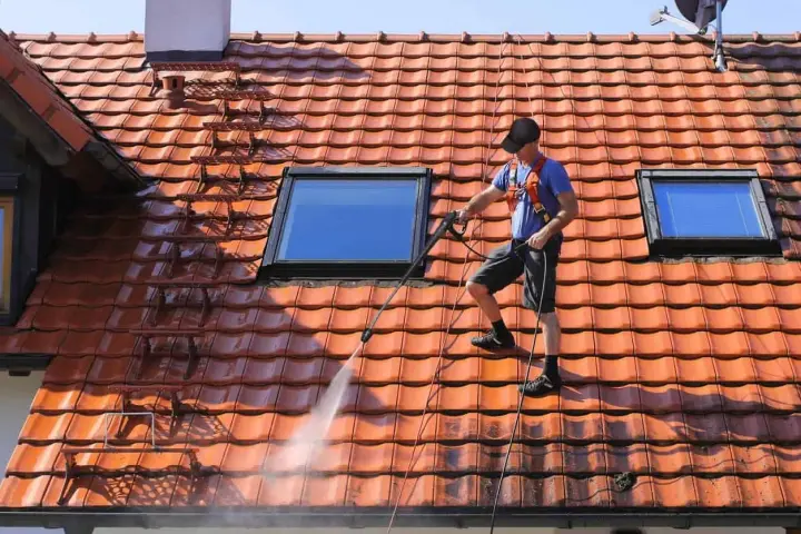 Un homme effectue le nettoyage toiture avec un nettoyeur haute pression sur un toit en tuiles rouges, sous un ciel bleu.