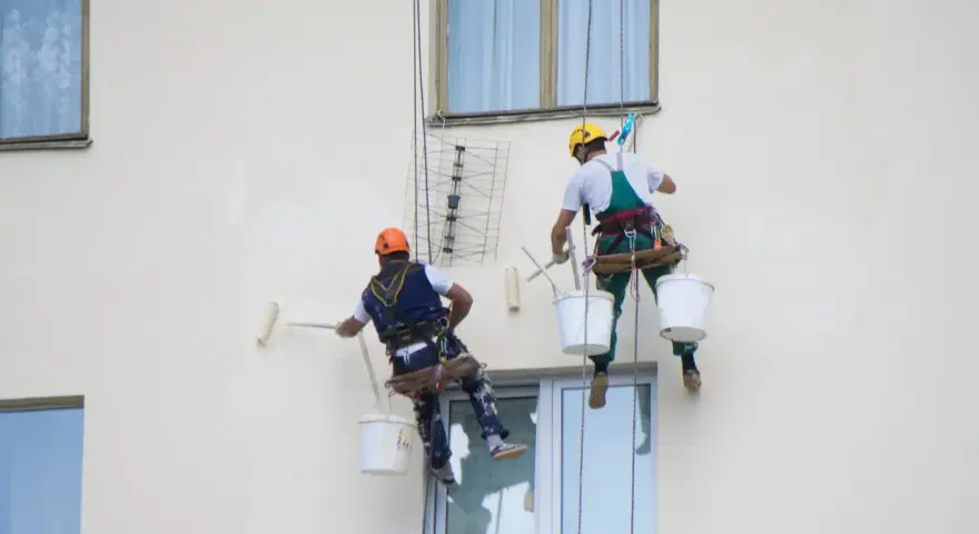 Deux peintres en bâtiment travaillent en hauteur sur une façade. L'un utilise un rouleau pour la peinture immeuble, l'autre est prêt à continuer.
