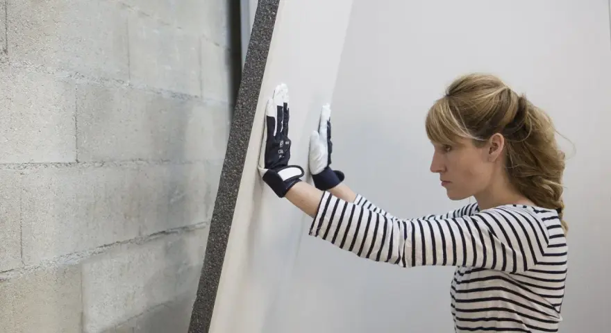 Une femme en gants manipule une plaque de doublage polystyrène expansé contre un mur de parpaings.