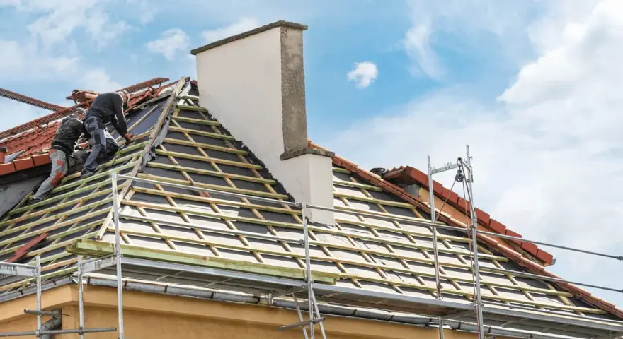 Deux couvreurs travaillent sur une toiture en cours de renovation. Des échafaudages entourent la maison.