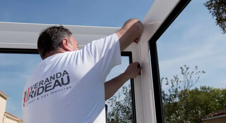 Pose d'un joint pour l'étanchéité véranda mur. Un ouvrier ajuste la structure blanche sous un ciel bleu.