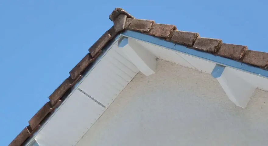 Le coin d'une toiture en pente avec des tuiles anciennes et un acrotère blanc.