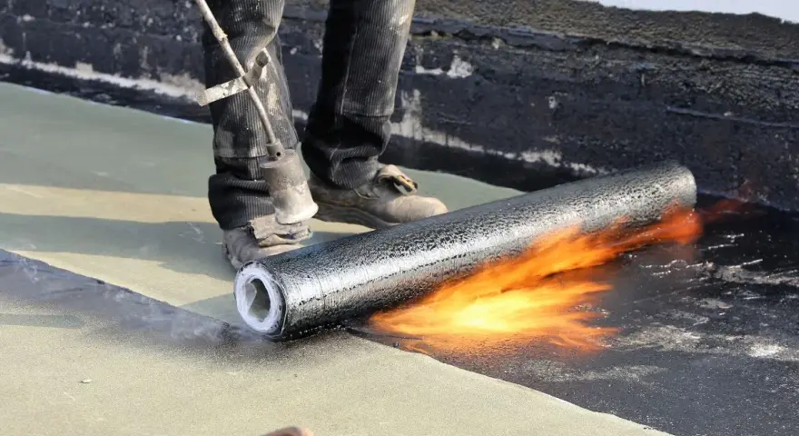 Un ouvrier réalise l'isolation d'un toit terrasse avec un chalumeau, fondant le revêtement pour une étanchéité parfaite.