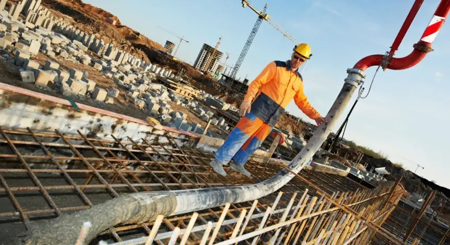 Un ouvrier en tenue orange et bleue coule du béton pour un mur banché sur un chantier. Grues et structures en construction en arrière-plan.