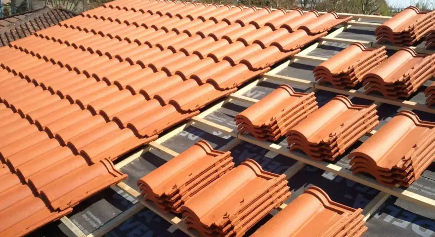 Vue d'un toit en cours de pose, révélant des rangées de tuiles mécaniques en terre cuite orange vif, empilées sur des lattes de bois.