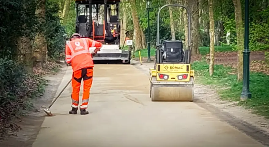 Un ouvrier prépare le chemin avec du sable stabilisé, tandis qu'un rouleau compresseur Bomag aplatit la surface.
