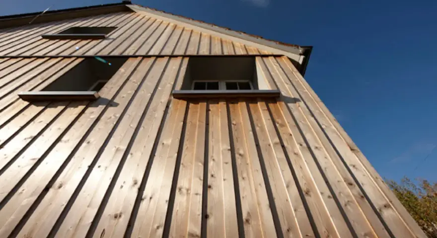 Vue en contre-plongée d'une maison avec un bardage bois clair, mettant en valeur la pose verticale des lames. Trois fenêtres sont visibles sous un ciel bleu éclatant.