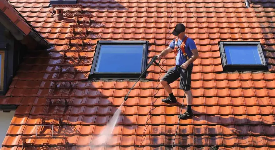 Un homme effectue le nettoyage toiture avec un nettoyeur haute pression sur un toit en tuiles rouges, sous un ciel bleu.