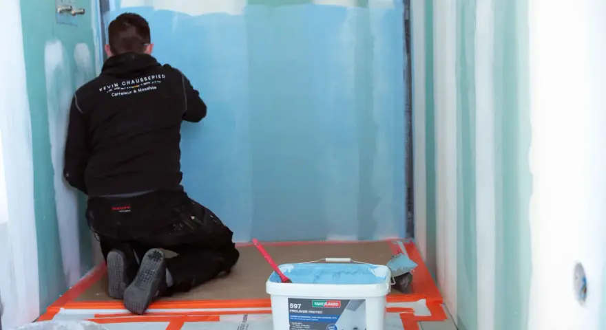 Un ouvrier applique un système d'étanchéité liquide bleu sur les murs d'une douche. Un seau de produit est visible au premier plan.