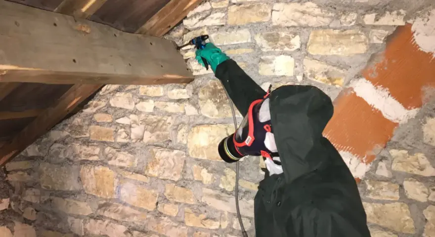 Personne en combinaison et masque effectuant un traitement de charpente par pulvérisation dans un grenier en pierre.