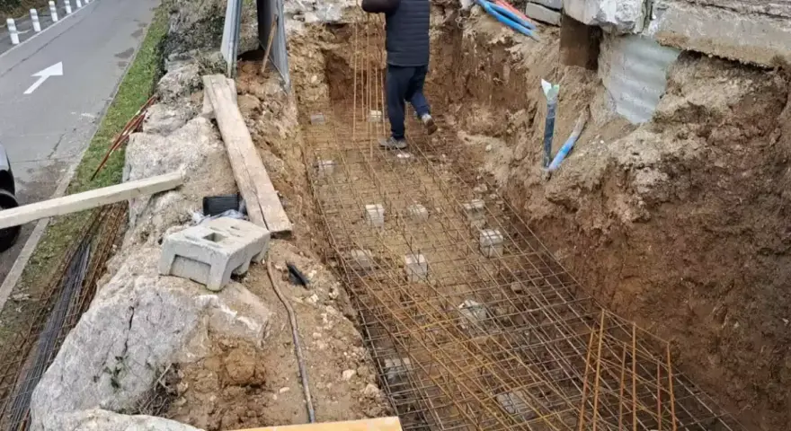 Un ouvrier travaille sur le ferraillage d'une fondation. Des armatures métalliques sont posées dans une fouille creusée dans la terre.
