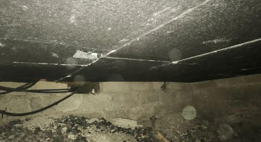 Vue de dessous d'un plancher, montrant le matériau d'isolation du plancher bas, des tuyaux et des blocs de béton.