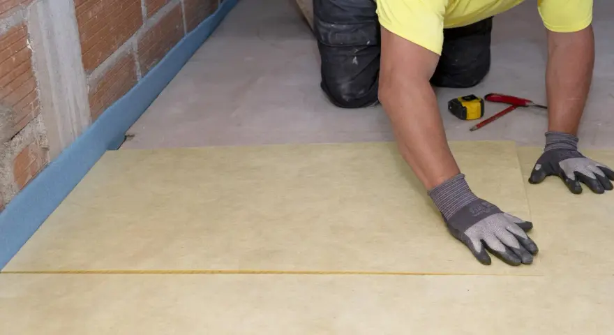 Pose d'une isolation sous dalle béton. Un ouvrier pose une feuille isolante jaune sur un sol brut, près d'un mur en briques.