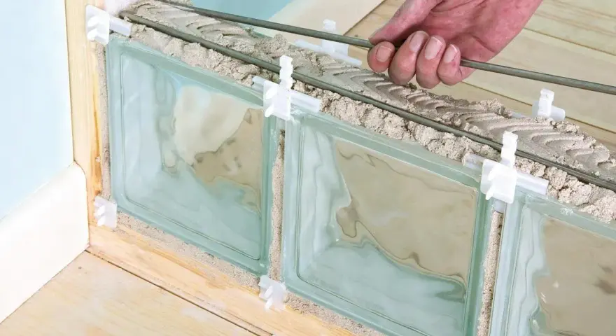 Pose de brique de verre extérieur : une main insère des barres de renfort dans le mortier entre les briques de verre, en cours d'installation.