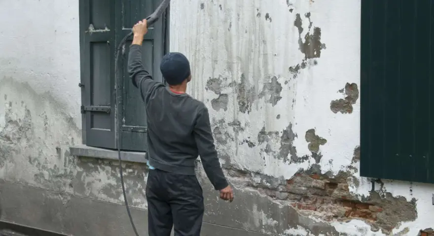 Un homme utilise un nettoyeur haute pression pour le sablage facade, révélant la brique sous l'enduit décrépit.