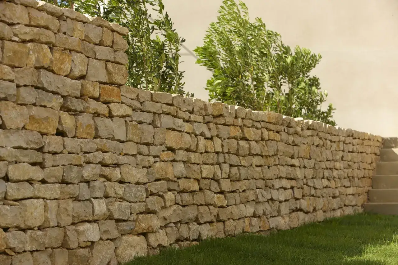 Un mur en pierre s&egrave;che, bord&eacute; d'herbe verte et d'arbustes luxuriants, s'&eacute;tend sur la droite, menant &agrave; des marches en pierre.