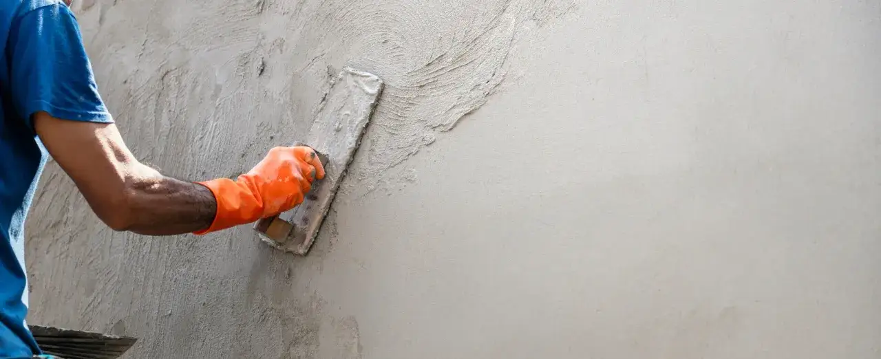 Main d'un ouvrier en gant orange appliquant de l'enduit sur agglo avec une truelle, cr&eacute;ant une texture sur le mur.