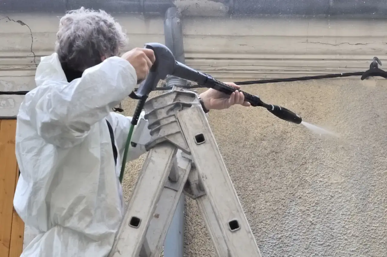 Person en combinaison blanche sur une échelle, utilisant un nettoyeur haute pression pour nettoyer une façade en crépi.