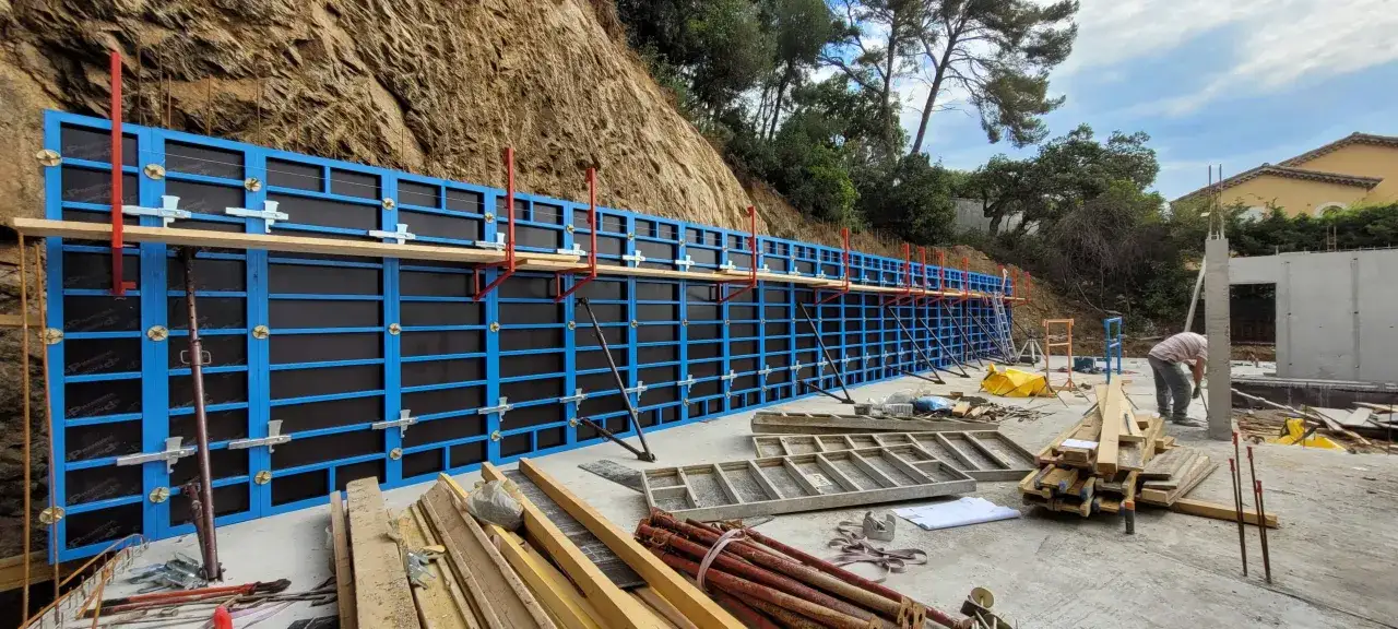 Un mur en b&eacute;ton est en cours de construction. Les ouvriers sont en train de faire un coffrage bleu et noir, soutenu par des &eacute;tais rouges et m&eacute;talliques, le long d'une colline rocheuse.