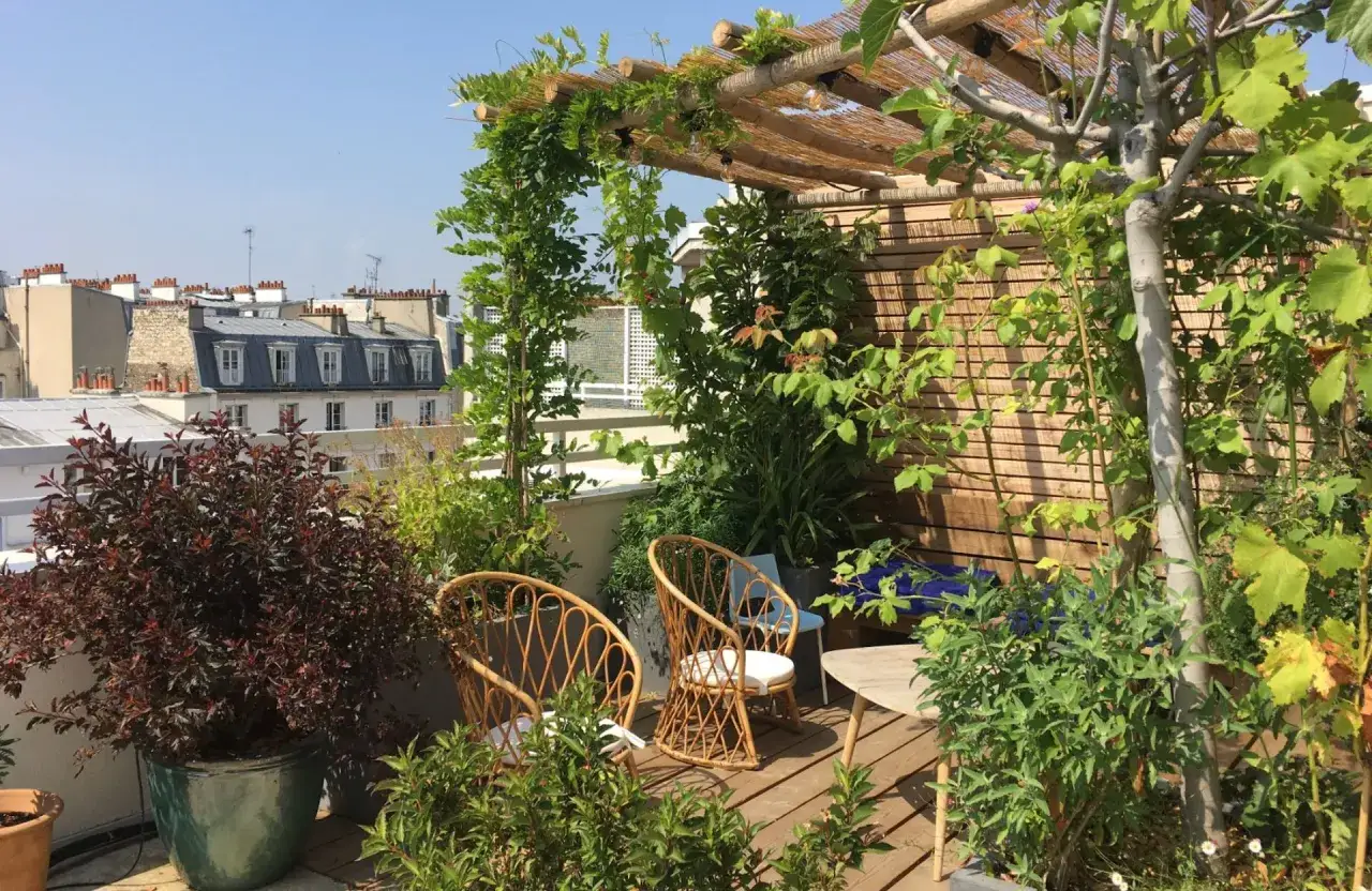 Am&eacute;nagement toit terrasse verdoyant avec chaises en rotin et pergola en bois. Vue sur les toits de Paris.