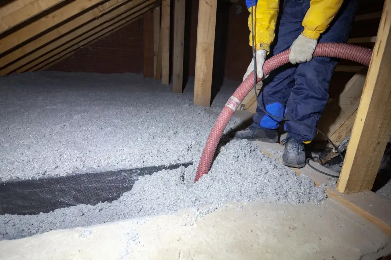 Un ouvrier installe de l'isolant en cellulose dans un grenier, soufflant le mat&eacute;riau par un tuyau rouge.
