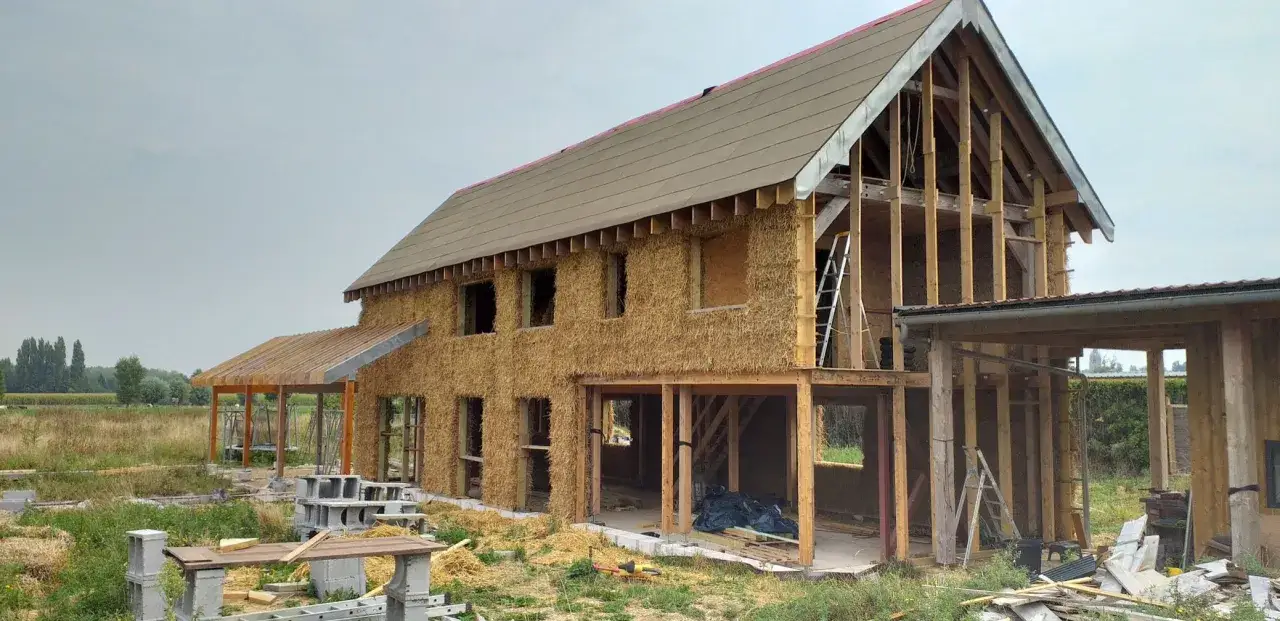 Maison en construction avec murs en bottes de paille, offrant une isolation naturelle. Le toit est en cours de finition.