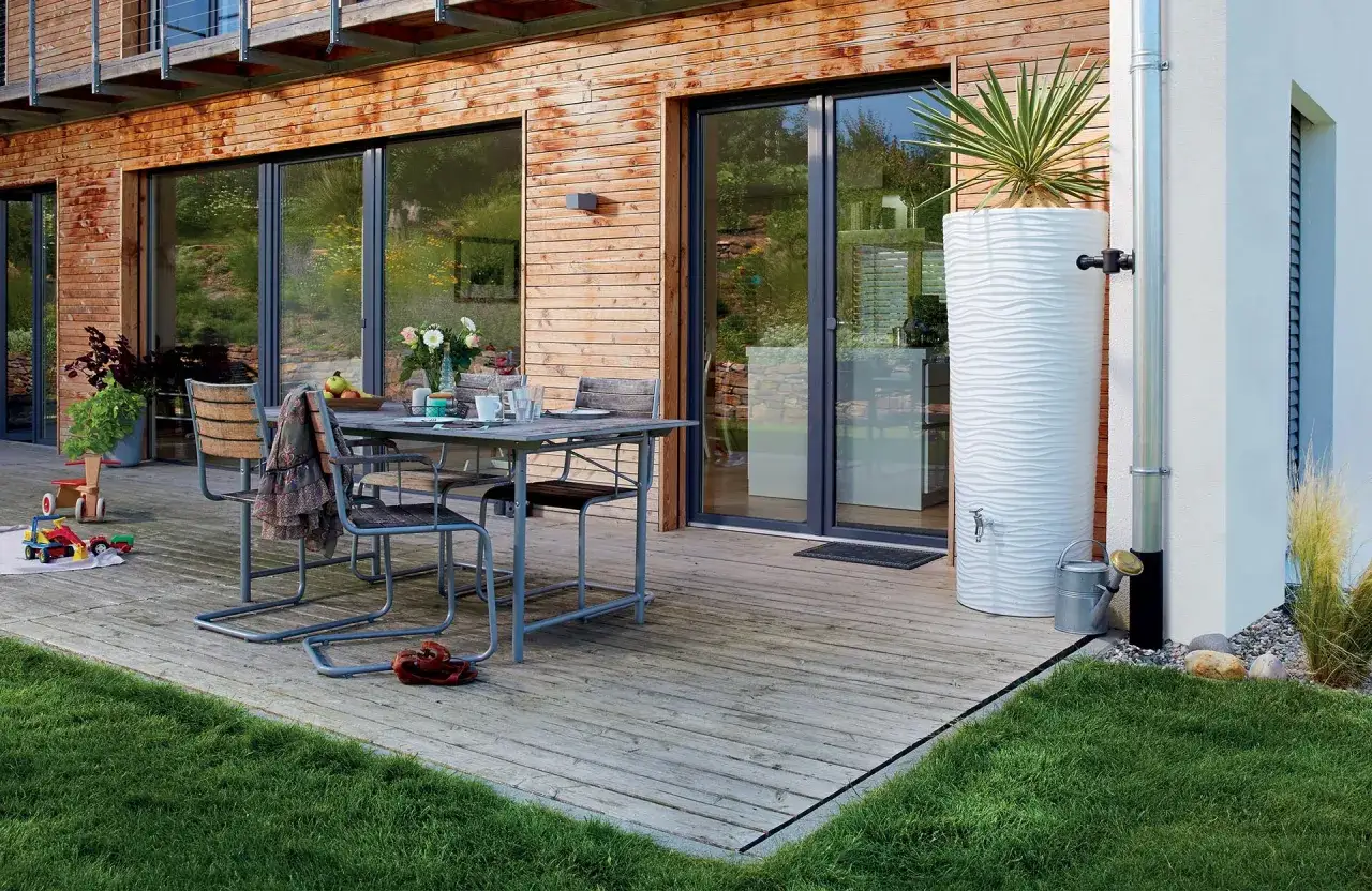 Un r&eacute;cup&eacute;rateur d'eau pluviale blanc, orn&eacute; d'une plante, est install&eacute; pr&egrave;s d'une maison en bois. Une goutti&egrave;re dirige l'eau vers le r&eacute;servoir.