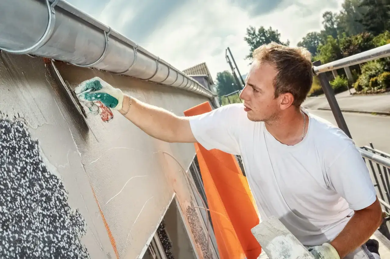 Un ouvrier applique un ragr&eacute;age sur un mur ext&eacute;rieur, utilisant une truelle pour lisser le mortier sur une armature.