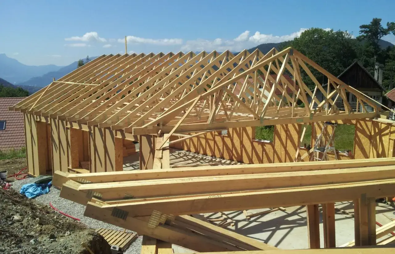 Construction d'une maison avec une charpente simple en bois, sous un ciel bleu. Les poutres s'étendent sur la structure en cours.