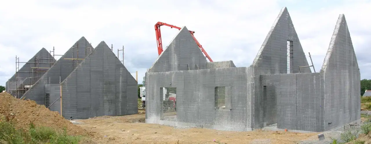 Chantier de maisons aux toits pointus en construction, utilisant un système de coffrage perdu en blocs de béton gris. Une grue rouge est visible.