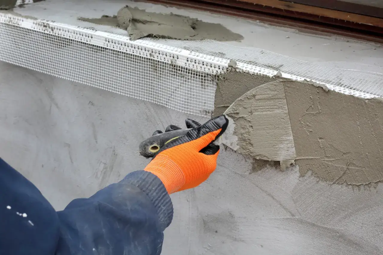 Application d'enduit sur une bande d'armature pour l'isolation sous enduit. Un ouvrier porte des gants orange et applique le mortier avec une truelle.