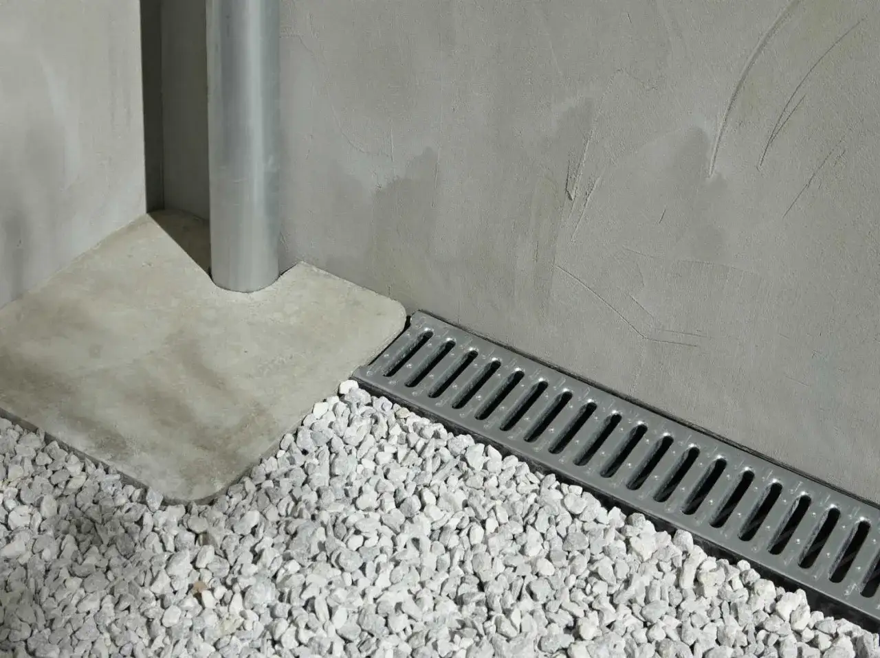 D&eacute;tail d'un pose caniveau en m&eacute;tal, bord&eacute; de gravier blanc, pr&egrave;s d'un mur en b&eacute;ton brut et d'une descente d'eau pluviale.
