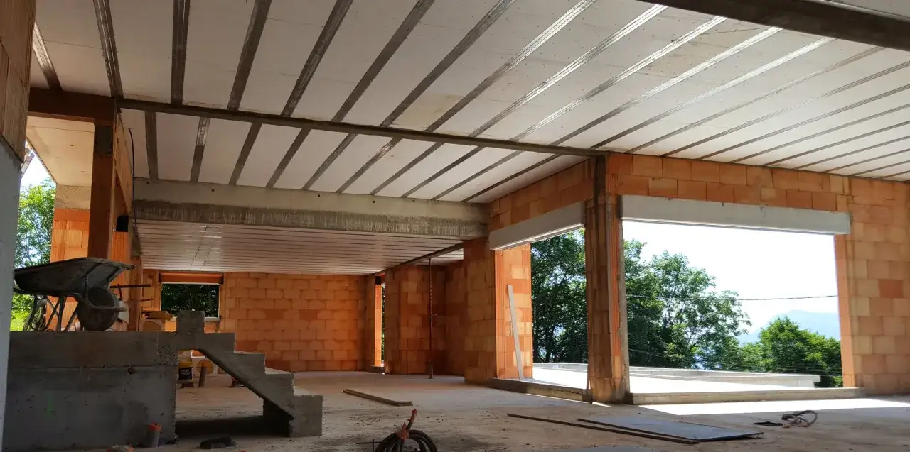 Intérieur d'une maison en construction, avec des murs en briques orange et un plafond composé d'hourdis béton. Une brouette et des escaliers en béton sont visibles.