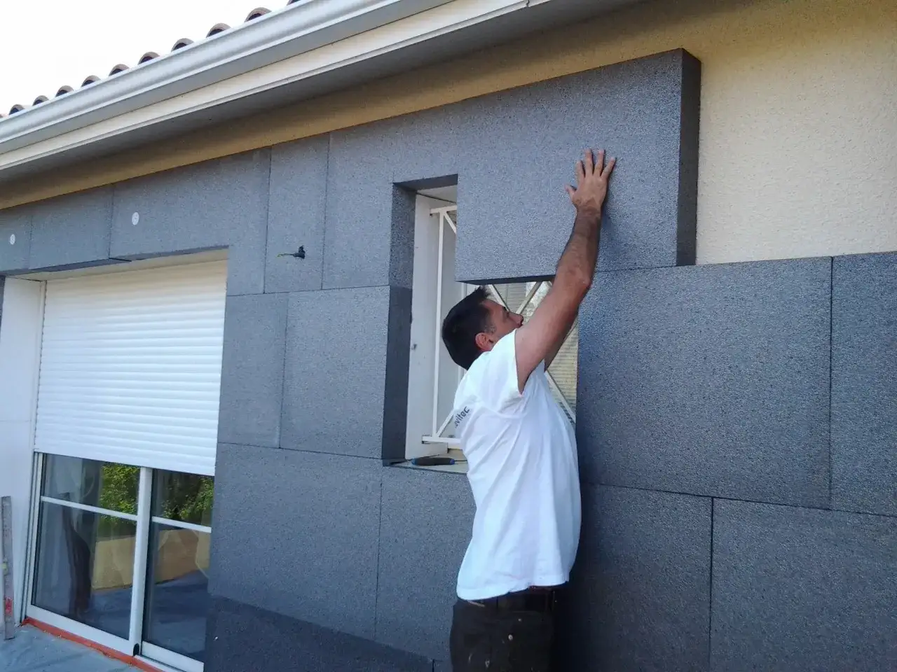 Un ouvrier pose des panneaux d'isolation polystyrène sur le mur extérieur d'une maison, près d'une fenêtre.