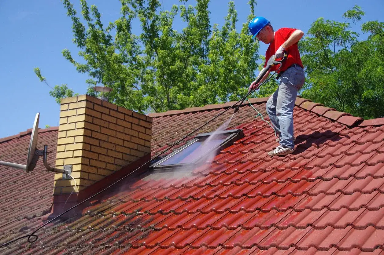 Un homme en casque bleu effectue un nettoyage toiture à haute pression sur un toit en tuiles rouges, à côté d'une cheminée en briques.