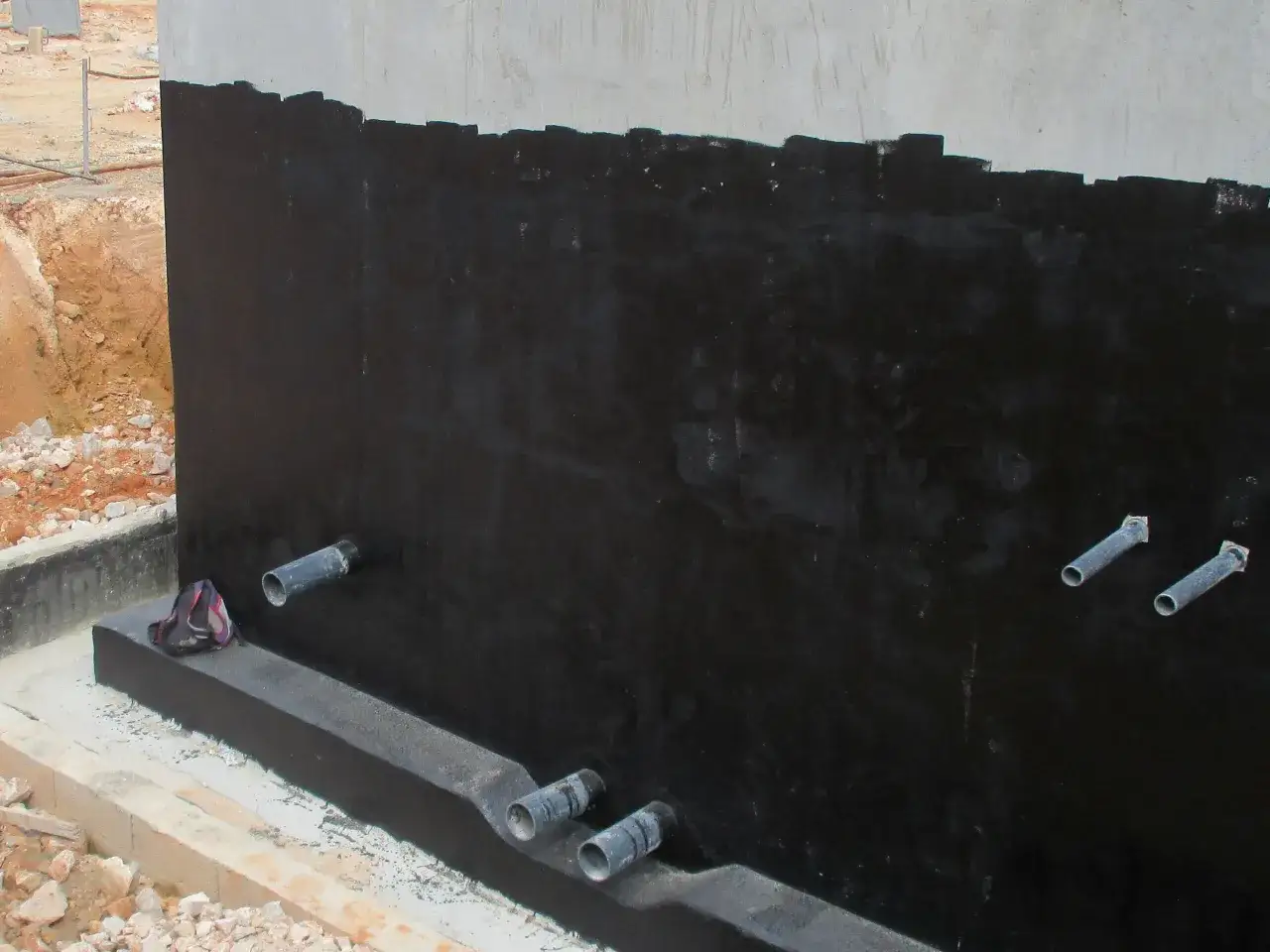 Application d'une membrane noire pour l'étanchéité d'un mur enterré. Des tuyaux sortent du mur, prêts pour le drainage.