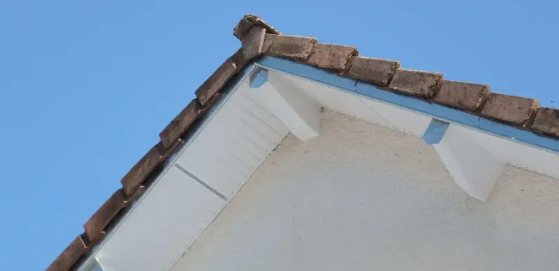 Le coin d'une toiture en pente avec des tuiles anciennes et un acrotère blanc.