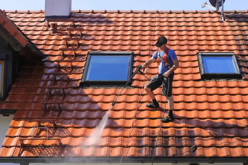 Un homme effectue le nettoyage toiture avec un nettoyeur haute pression sur un toit en tuiles rouges, sous un ciel bleu.
