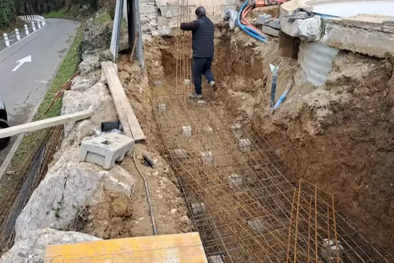 Un ouvrier travaille sur le ferraillage d'une fondation. Des armatures métalliques sont posées dans une fouille creusée dans la terre.