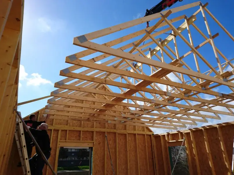 Construction d'une charpente fermette en bois sous un ciel bleu. Des ouvriers travaillent sur l'installation.